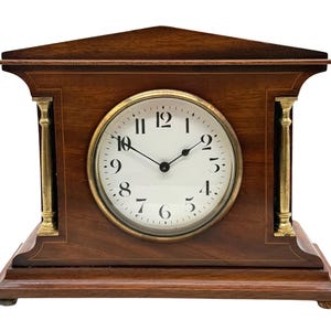 Puede incluir: Un reloj de sobremesa de madera con acabado marrón oscuro, que presenta una parte superior triangular, una esfera circular blanca con números arábigos negros y dos manecillas negras finas. El reloj incluye dos columnas verticales de latón.