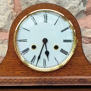 May include: A wooden mantel clock with a white face and black Roman numeral hour markers. The clock has gold-coloured hands and a glass cover.