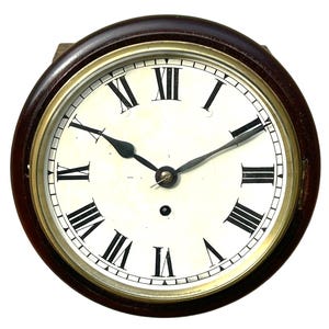 Puede incluir: Un reloj de pared redondo con un marco de madera marrón oscuro y un bisel de latón. La esfera de color blanco roto muestra números romanos negros y dos manecillas de metal negro. Un pequeño orificio de cuerda negro se sitúa bajo el centro.