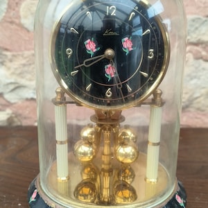 May include: A vintage clock in a glass dome. The black clock face has gold hands, numerals, and pink rose details. The base is black with gold trim and rose accents. The clock features a pendulum with gold spheres.