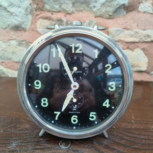 May include: A silver alarm clock with a black face and white numbers. The clock has a repetition feature and the text "JAZ EMPESTO" is printed on the face.