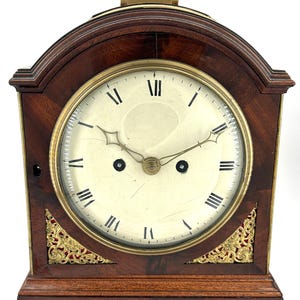 Könnte beinhalten: Eine antike hölzerne Kaminuhr mit cremefarbenem Zifferblatt und römischen Ziffern. Die Uhr hat einen Messinggriff und -verzierungen mit dekorativen goldenen Eckakzenten. Das dunkle Holzgehäuse ist rechteckig mit gewölbter Oberseite.
