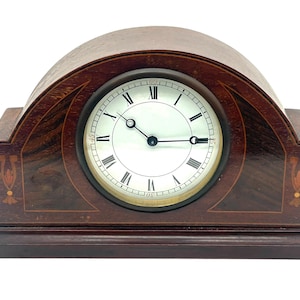 Puede incluir: Un reloj de sobremesa de madera vintage con esfera blanca y números romanos negros. El reloj tiene un acabado marrón oscuro con patrones incrustados decorativos y una parte superior curvada. Las manecillas del reloj señalan las 2:15.