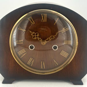 Puede incluir: Un reloj de chimenea de madera vintage con una esfera dorada y marcadores de hora en números romanos. El reloj tiene una cubierta de vidrio y una caja de madera oscura.