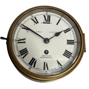 Puede incluir: Un reloj de barco de latón con números romanos y una esfera blanca. La esfera del reloj dice "H.M.S. HOOD" y "SMITH'S CRICKLEWOOD. N.W.2 MADE IN ENGLAND".