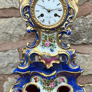 Puede incluir: Un reloj de chimenea de porcelana azul y dorado con una esfera blanca y agujas negras. El reloj está decorado con detalles florales y tiene una base dorada.