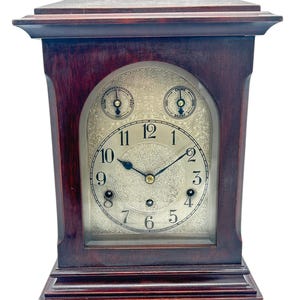 Puede incluir: Un reloj de chimenea de madera de caoba con una esfera plateada y agujas negras. El reloj tiene un ajuste de timbre y silencio.