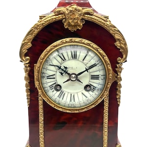 Op de afbeelding: Een antieke mantelklok met een bordeauxrode afwerking en goudkleurige accenten. De klok heeft Romeinse cijfers en zwarte wijzers. De klok heeft sierlijke details, waaronder een decoratieve boven- en onderkant.
