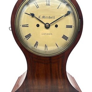 Puede incluir: Un reloj de sobremesa de madera vintage con esfera color crema y números romanos. La esfera del reloj dice "C. Mitchell LONDON". El reloj tiene una caja de madera marrón oscuro con la parte superior e inferior curvadas y detalles en latón. El reloj mide unos 25 cm de alto.
