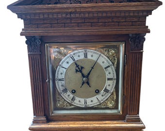 May include: Antique wooden mantel clock with a Roman numeral clock face and a carved wooden top. The clock has a brick-like design on the base and sides.
