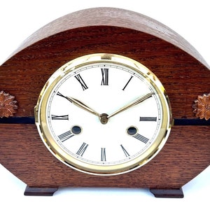 May include: A wooden mantel clock with a white clock face and gold-coloured trim. The clock has Roman numerals and decorative floral carvings on the sides. The clock is approximately 25 cm wide.