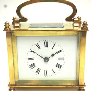 May include: A brass clock with a white face and black Roman numeral hour markers. The clock has a handle on the top and sits on four small feet.