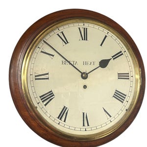 Könnte beinhalten: Eine runde Uhr mit einem Holzrahmen und einem cremefarbenen Zifferblatt. Die Uhr hat schwarze römische Ziffern und Zeiger, mit dem Text "BETTA HEAT" in der Mitte. Sie hat einen goldfarbenen Innenring.