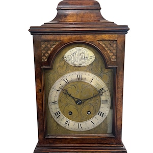 May include: A wooden antique clock with a decorative handle. The clock face features Roman numerals and ornate detailing. A ship illustration is above the clock face. The clock is brown and has a rectangular shape.