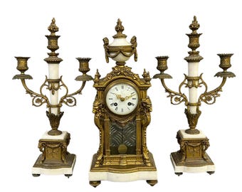 Könnte beinhalten: Ein Set aus drei antiken goldfarbenen Kerzenständern und einer Uhr. Die Uhr hat ein weißes Zifferblatt mit schwarzen römischen Ziffern. Die Kerzenleuchter haben weiße Marmorsockel und goldfarbene Akzente.