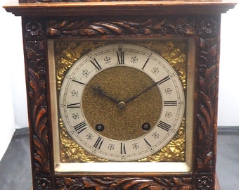 May include: A dark brown antique clock with a square face and ornate carved wooden frame. The clock face has Roman numerals and a gold-colored center. The clock is a decorative piece.