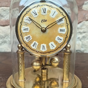 May include: A gold-toned pendulum clock with Roman numeral markings on the face. The clock is encased in a clear glass dome and sits on a gold-toned base.