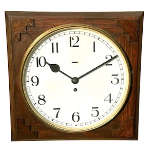 May include: A square wall clock with a dark brown wooden frame featuring stepped corner details. The clock face is white with black Arabic numerals from 1 to 12, black hour and minute hands, and a circular brass-toned bezel surrounding the dial.
