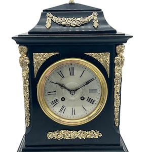 May include: A black and gold mantel clock with a Roman numeral clock face. The clock has ornate gold accents and a decorative top.
