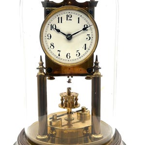 May include: A brass anniversary clock with a white circular face, black Arabic numerals, and black hands, enclosed within a clear glass dome. The clock features a rotating pendulum mechanism at the base, supported by four brass pillars on a round base.