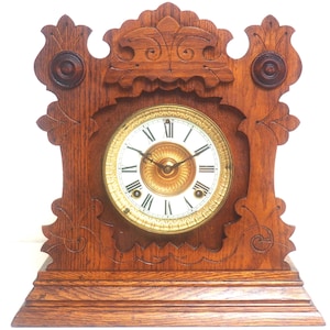 May include: A vintage wooden mantel clock with a light brown finish. The clock face is white with black Roman numerals and gold-colored hands and trim. The clock has decorative carvings and a detailed design.