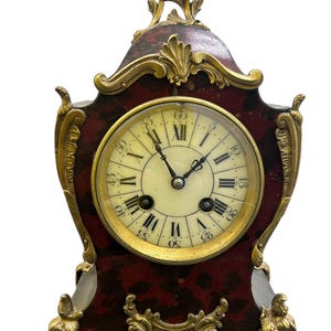 Puede incluir: Un reloj antiguo, adornado, con detalles dorados y números romanos en una esfera blanca. El reloj tiene una base de mármol rojo y negro con detalles dorados y patas ornamentadas.