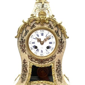 May include: A mantel clock featuring a cream-colored body with intricate gold-toned metal filigree accents. The clock face has black Roman numerals and ornate hands. A gold-toned cherub figure sits atop the clock, and a dark glass panel is below.