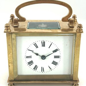Puede incluir: Un reloj de latón con una esfera blanca y marcadores de hora con números romanos. El reloj tiene una cubierta de vidrio y una manija en la parte superior.
