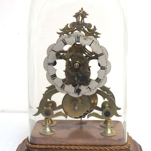 May include: A brass clock with a white face and Roman numerals, encased in a glass dome. The clock sits on a wooden base with a rope trim.