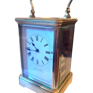 May include: A rectangular brass carriage clock with a white clock face featuring black Roman numerals and hands. The clock has a brass handle and is encased in glass panels. The clock is a vintage item.