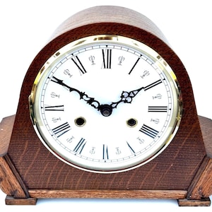 May include: A dark brown wooden mantel clock with a white face and black Roman numerals. The clock has a gold-coloured bezel and black hands. The clock's wooden case is a dark brown colour.