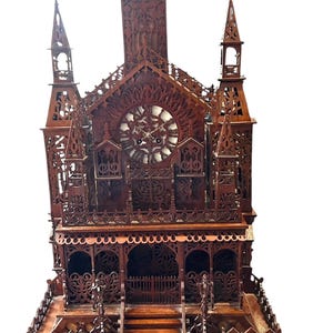 Peut inclure: Une horloge en bois brun foncé en forme de cathédrale avec des découpes complexes, plusieurs flèches et un cadran circulaire central. La structure présente des détails ajourés, une base à gradins et une balustrade décorative à l'avant.