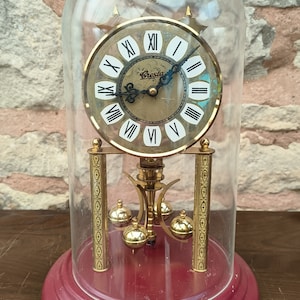 May include: A vintage clock under a glass dome. The clock face is gold with black Roman numerals and the brand name "Cresta". The base is a deep red, and the clock has gold pendulum and decorative elements.