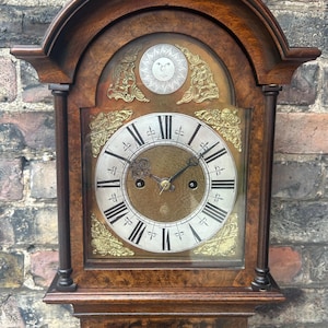 Puede incluir: Un reloj de pie antiguo y alto con una caja de madera y una esfera blanca. El reloj tiene una esfera con números romanos y un diseño de sol y luna sobre la esfera.