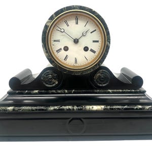 May include: A mantel clock featuring a circular face with black Roman numerals and ornate hands, encased in a dark green and black marbled stone frame. The base is constructed from smooth black material with a rectangular panel and circular detail.