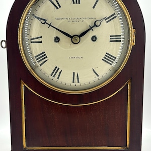 Puede incluir: Un reloj de chimenea de caoba con una esfera de latón y detalles dorados. La esfera del reloj dice "Goldsmiths & Silversmiths Company 112, Regent St. London".