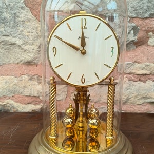 May include: A vintage gold-toned clock with a white face, encased in a clear glass dome. The clock features gold hands and a pendulum with multiple gold spheres. The base is circular, and the clock has the word "Bema" on the face.