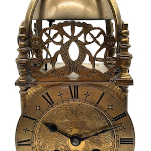 May include: An antique brass clock with Roman numerals on the face. The clock has an ornate design with a domed top and decorative embellishments. The clock's face features intricate floral patterns and is supported by four decorative feet.