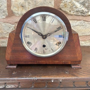 May include: A vintage wooden mantel clock with a silver face and Roman numeral hour markers. The clock has a rounded top and a rectangular base.