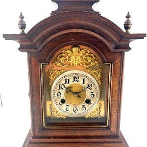 Puede incluir: Un reloj de chimenea de madera oscura con una esfera dorada y detalles ornamentados. El reloj tiene una esfera romana con manecillas negras.