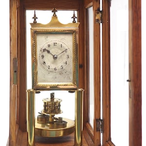 May include: A vintage brass clock with a white face and black hands, housed in a wooden case with a glass door. The clock is on a wooden base and has a pendulum.