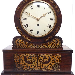 May include: A vintage wooden mantel clock with a round, white face and black Roman numeral hour markers. The clock has a decorative, inlaid wood design with gold accents.