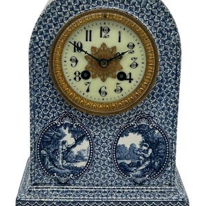Puede incluir: Un reloj de chimenea de porcelana azul y blanco con una esfera dorada y marcadores de hora en números romanos. El reloj presenta dos escenas ovaladas de personas en un paisaje.