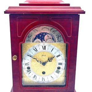 Puede incluir: Un reloj de chimenea de madera de caoba con una esfera dorada y marcadores de hora con números romanos. El reloj tiene un dial de fase lunar con una representación de la luna y las estrellas. La esfera del reloj dice "Hermle".