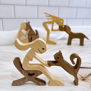 Puede incluir: Figuras de madera de una mujer, un gato, un perro y una persona con piernas largas. La mujer está sentada en una silla de madera y sostiene la pata del gato. El gato es marrón y la mujer es marrón claro. El perro es marrón y la persona con piernas largas es marrón claro.