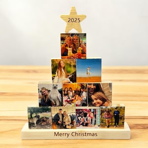 Könnte beinhalten: Ein weihnachtsbaumförmiges Display aus Holz mit Fotos auf Holzblöcken. Ein Holzstern mit der Aufschrift "2025" sitzt oben auf dem Baum. Die Basis trägt die Aufschrift "Merry Christmas". Die Fotos zeigen Menschen und Szenen.