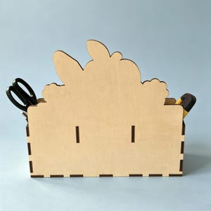 Op de afbeelding: Een bureau-organizer van lichtgekleurd hout met een decoratieve uitgesneden vorm die lijkt op een konijn met lange oren. De rechthoekige houder heeft zichtbare in elkaar grijpende verbindingen langs de randen en bevat een zwarte schaar links.