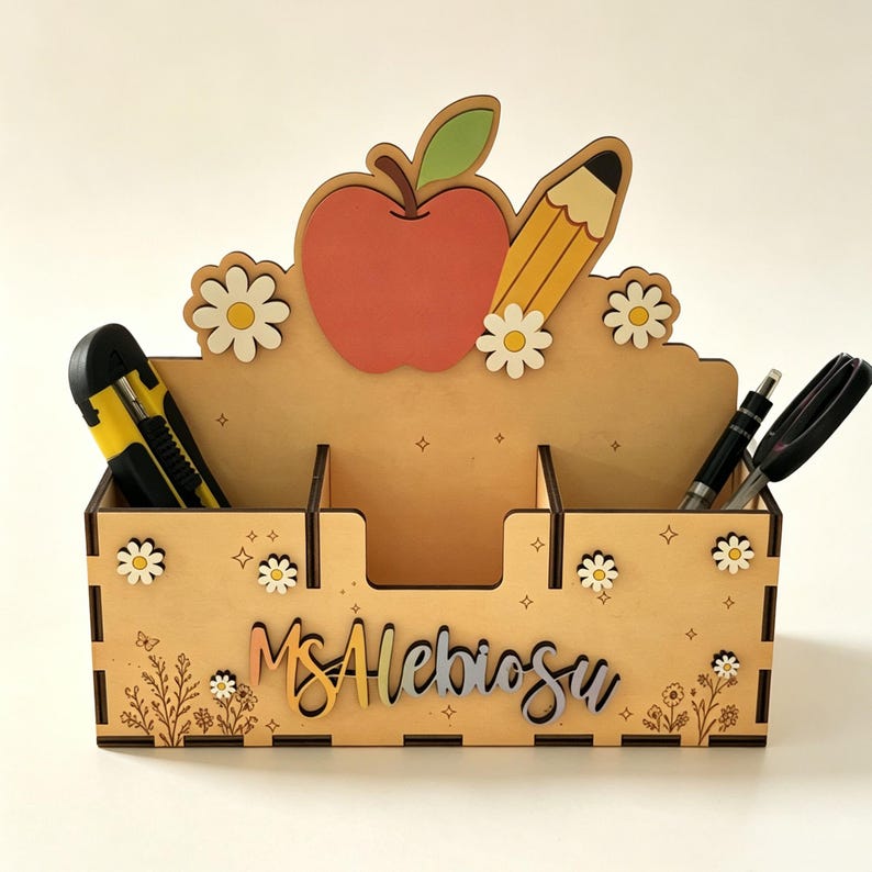Op de afbeelding: Een houten bureau-organizer met een uitgesneden rode appel en een geel potlood aan de bovenkant. Het voorpaneel is versierd met witte madeliefjes, kleine sterretjes en bloemmotieven. De naam Ms. Alebiosu is weergegeven in een cursief lettertype.