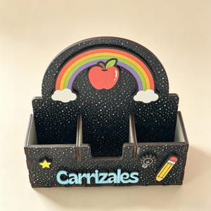 Op de afbeelding: Een zwarte bureau-organizer met een sterrenpatroon, voorzien van een regenboog en een rode appel op het achterpaneel. De voorzijde toont de naam Carrizales in lichtblauwe letters, een gele ster, een wit gloeilampicoon en een geel potlood.