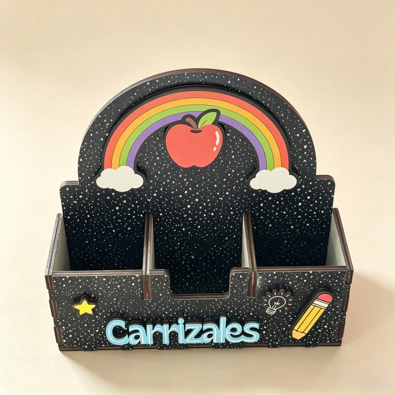 Op de afbeelding: Een zwarte bureau-organizer met een sterrenpatroon, voorzien van een regenboog en een rode appel op het achterpaneel. De voorzijde toont de naam Carrizales in lichtblauwe letters, een gele ster, een wit gloeilampicoon en een geel potlood.